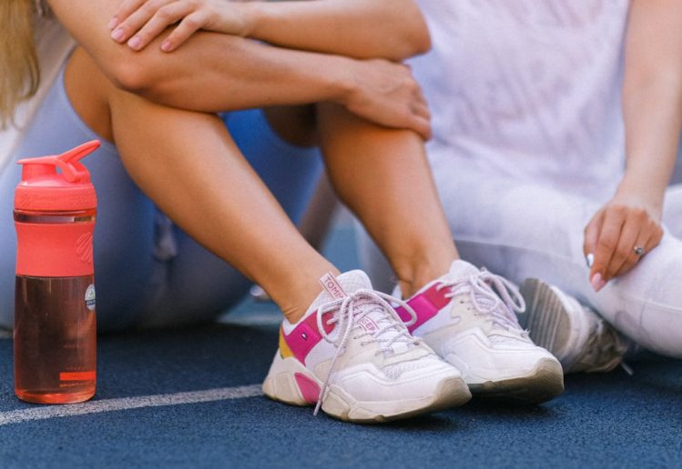 Crop anonymous female friends in activewear sitting on sports ground after training near bottle with water in daytime