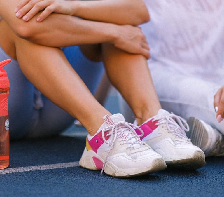 Crop anonymous female friends in activewear sitting on sports ground after training near bottle with water in daytime